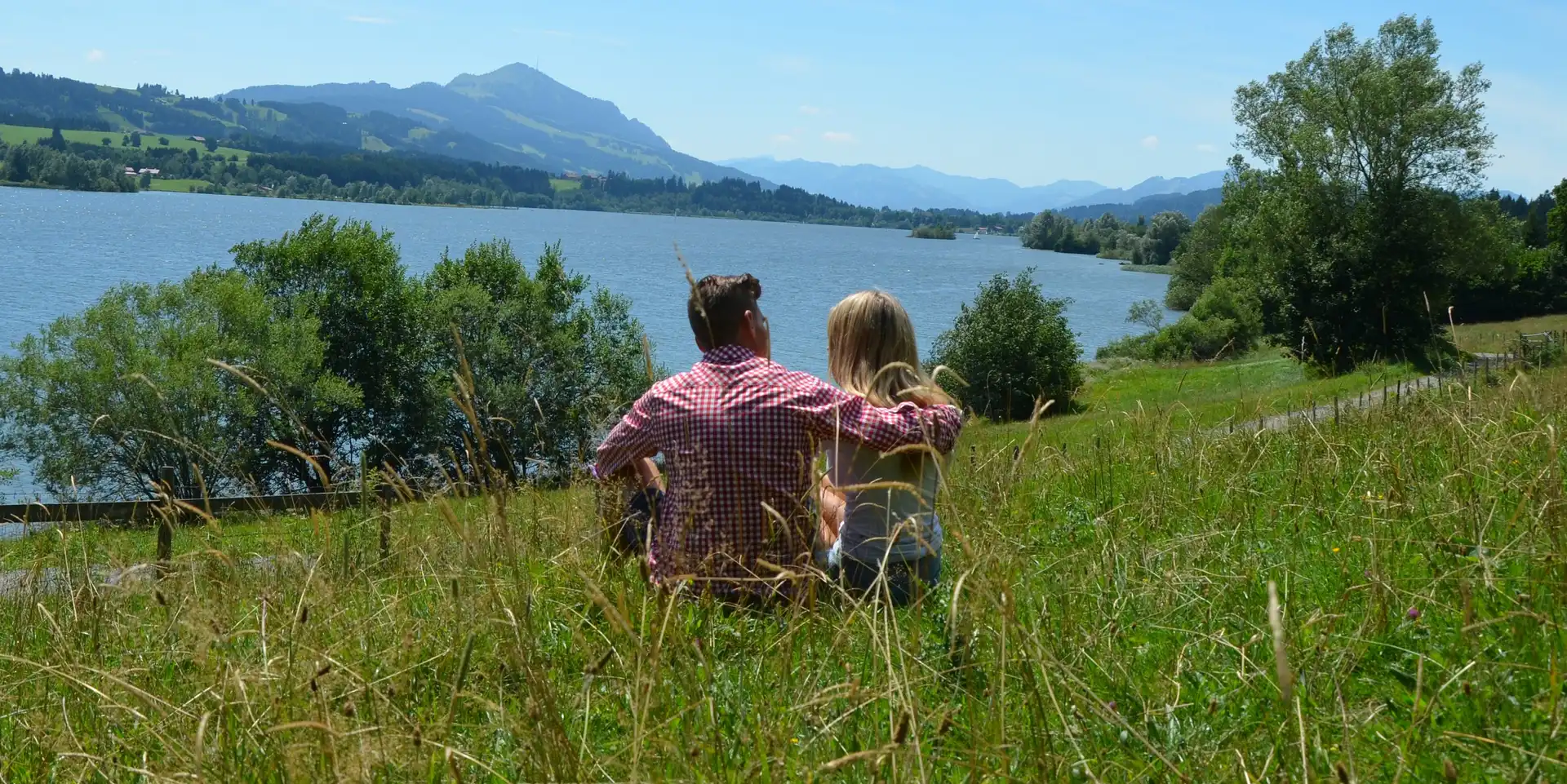 Ein Paar sitzt auf einer Wiese am Ufer eines Sees. Im Hintergrund sind Hügel und Berge sichtbar, und der Himmel ist klar und blau. Das Paar blickt gemeinsam auf die Landschaft und geniesst die Natur.