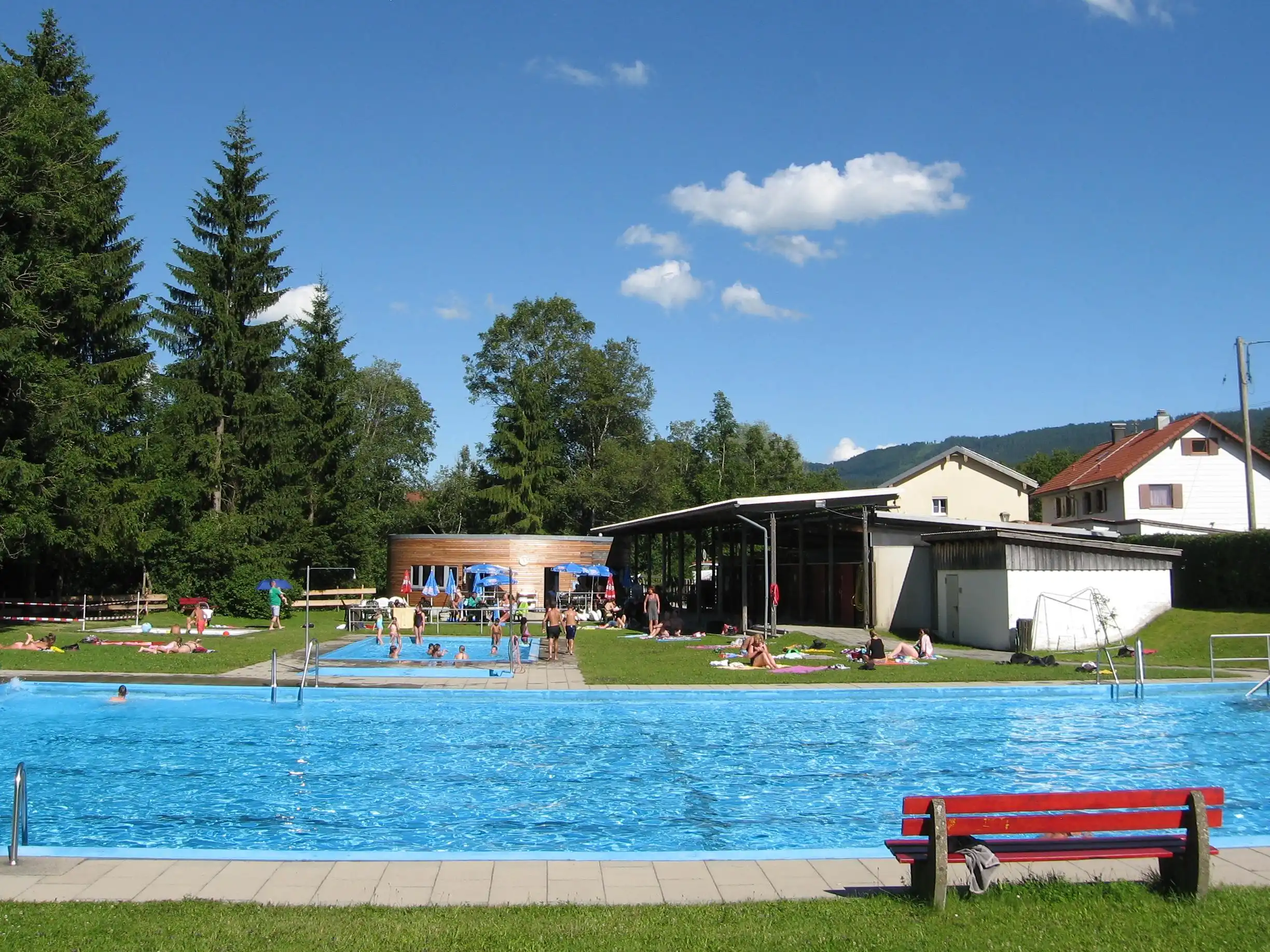 Mehrere Personen genießen ein Schwimmbad im Freien, umgeben von grüner Landschaft und Bäumen. Im Hintergrund sind Gebäude und eine Schwimmhalle sichtbar. Es ist sonnig und es gibt Liegen und Bänke im Bereich des Pools.
